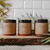 Herbal Bath Salts in amber jars. There is a relaxing bath salt, reviving bath salt, and seduction bath salt. They are placed on a block of wood in front of subway tiles and a palm tree.
