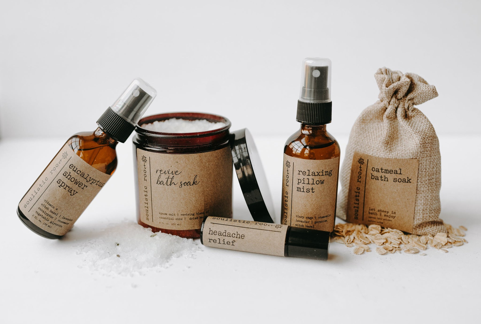The spa gift set which includes a eucalyptus shower spray, organic lavender oatmeal bath soak, headache relief roller, essential oil & herb revive bath soak and a relaxing pillow mist. The set is on a blanket in front of a white background with some of the oatmeal and epsom salt & herbs spilt out.