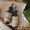 An essential oil mask spray and moisturizing hand sanitizer next to each other on a block of wood with a plant in the background.