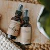 Essential oil mask spray and moisturizing hand sanitizer next to each other on a wooden block next to a knit blanket and plant. They are both in 2 oz amber spray bottles.