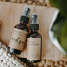 All natural hand sanitizer and an essential oil mask spray next to each other on a wood block with a blanket and plant in the background.
