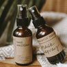 Essential oil mask spray and moisturizing hand sanitizer next to each other on a grey blanket with a palm leaf in the background. They are both in 2 oz amber spray bottles.