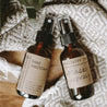 Essential oil mask spray and moisturizing hand sanitizer next to each other on a grey blanket. They are in 2 oz amber bottles.