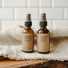 Makeup setting spray and a eucalyptus shower spray on a piece of wood with a white blanket in front of subway tiles. Both products are in a 2 oz amber glass spray bottle.