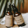 An essential oil mask spray and moisturizing hand sanitizer next to each other on a block of wood and grey and white blanket with a plant in the background.