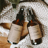 An essential oil mask spray and moisturizing hand sanitizer next to each other on a block of wood and grey and white blanket.