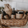 The spa gift set which includes a eucalyptus shower spray, organic lavender oatmeal bath soak, headache relief roller, essential oil & herb revive bath soak and a relaxing pillow mist. The set is on a blanket inside of a basket in front of subway tile.