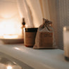 A lifestyle shot of an organic herbal oatmeal bath soak in a burlap bag, a relaxing epsom salt bath with lavender and chamomile, and a shower spray all in a bath tub full of water. There is also a lit candle.