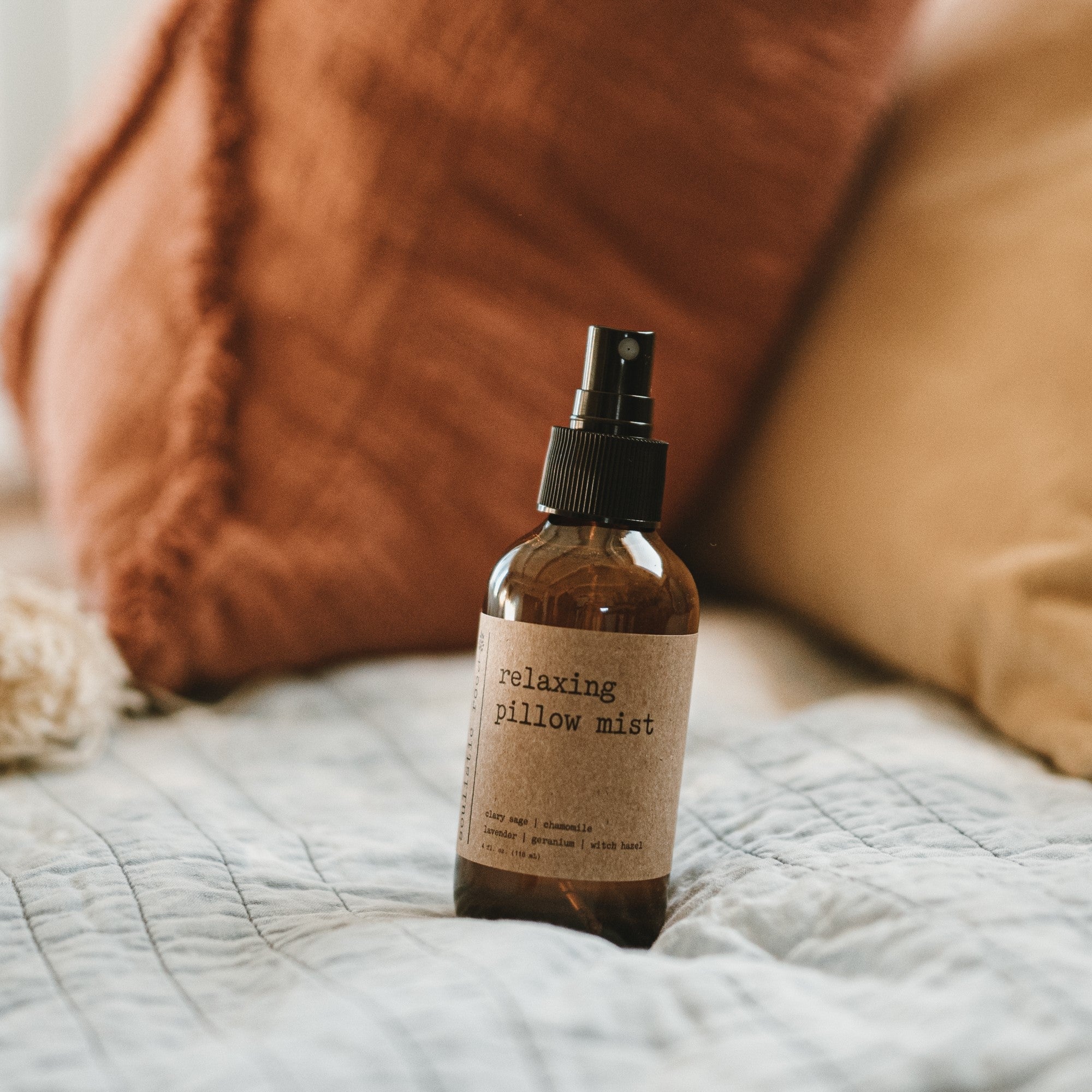Relaxing pillow mist spray in an amber glass bottle on a bed with burnt orange pillows.