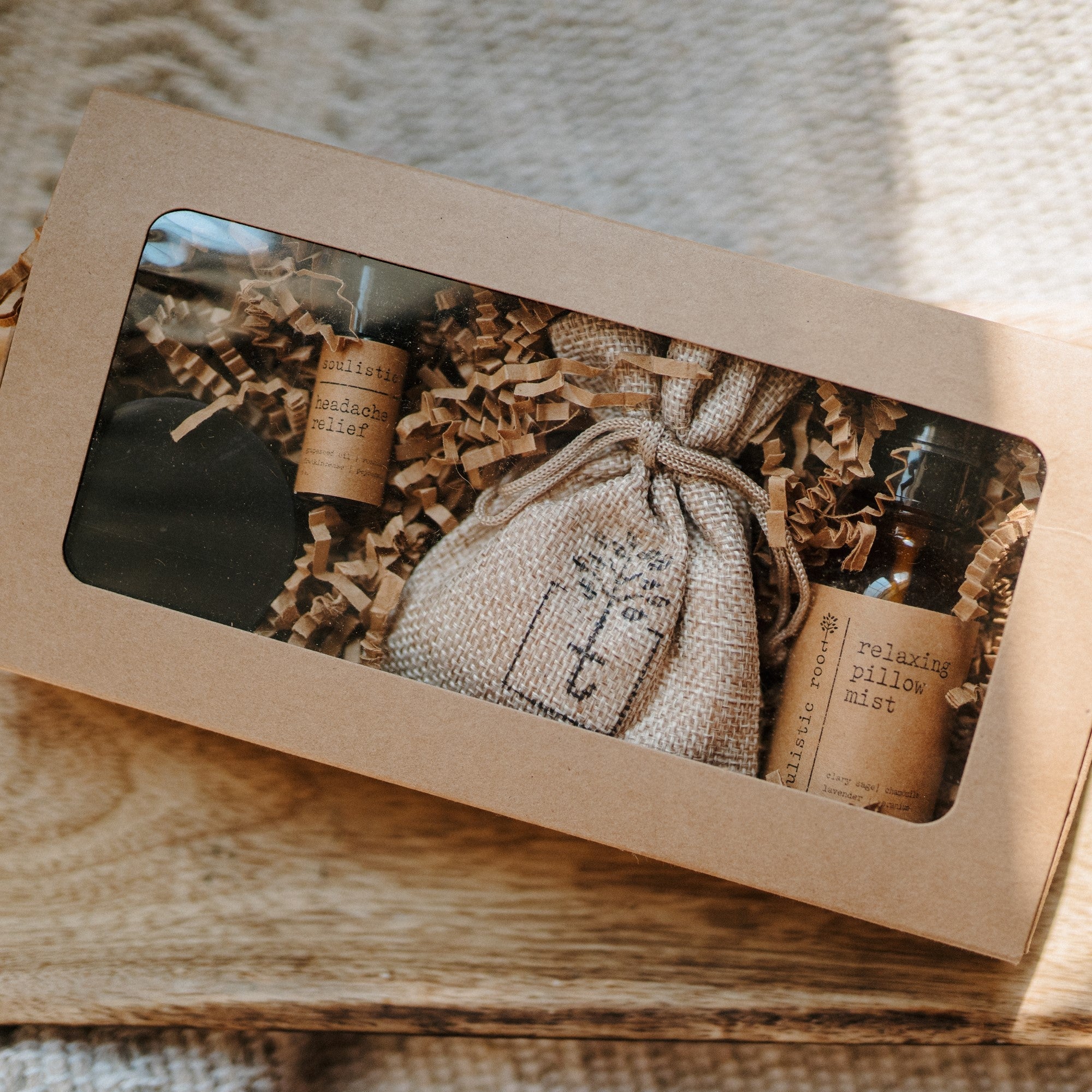 A travel set full of sample sizes already wrapped in a kraft box. The set includes a lavender & chamomile epsom salt bath, essential oil headache relief roller, organic herbal oatmeal bath soak & a relaxing pillow spray. The set is on a piece of wood on a white blanket.