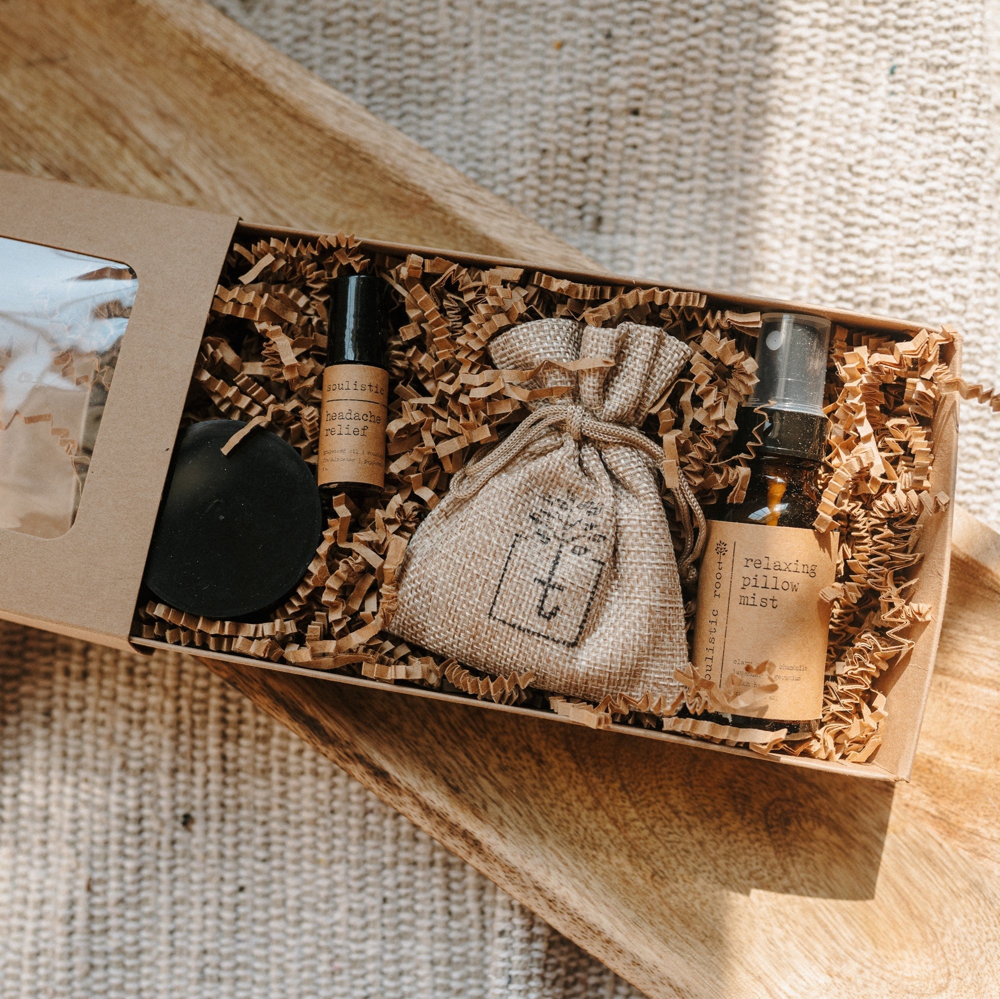 A travel set full of sample sizes already wrapped in a kraft box. The set includes a lavender & chamomile epsom salt bath, essential oil headache relief roller, organic herbal oatmeal bath soak & a relaxing pillow spray. The set is on a piece of wood and blanket in the background.