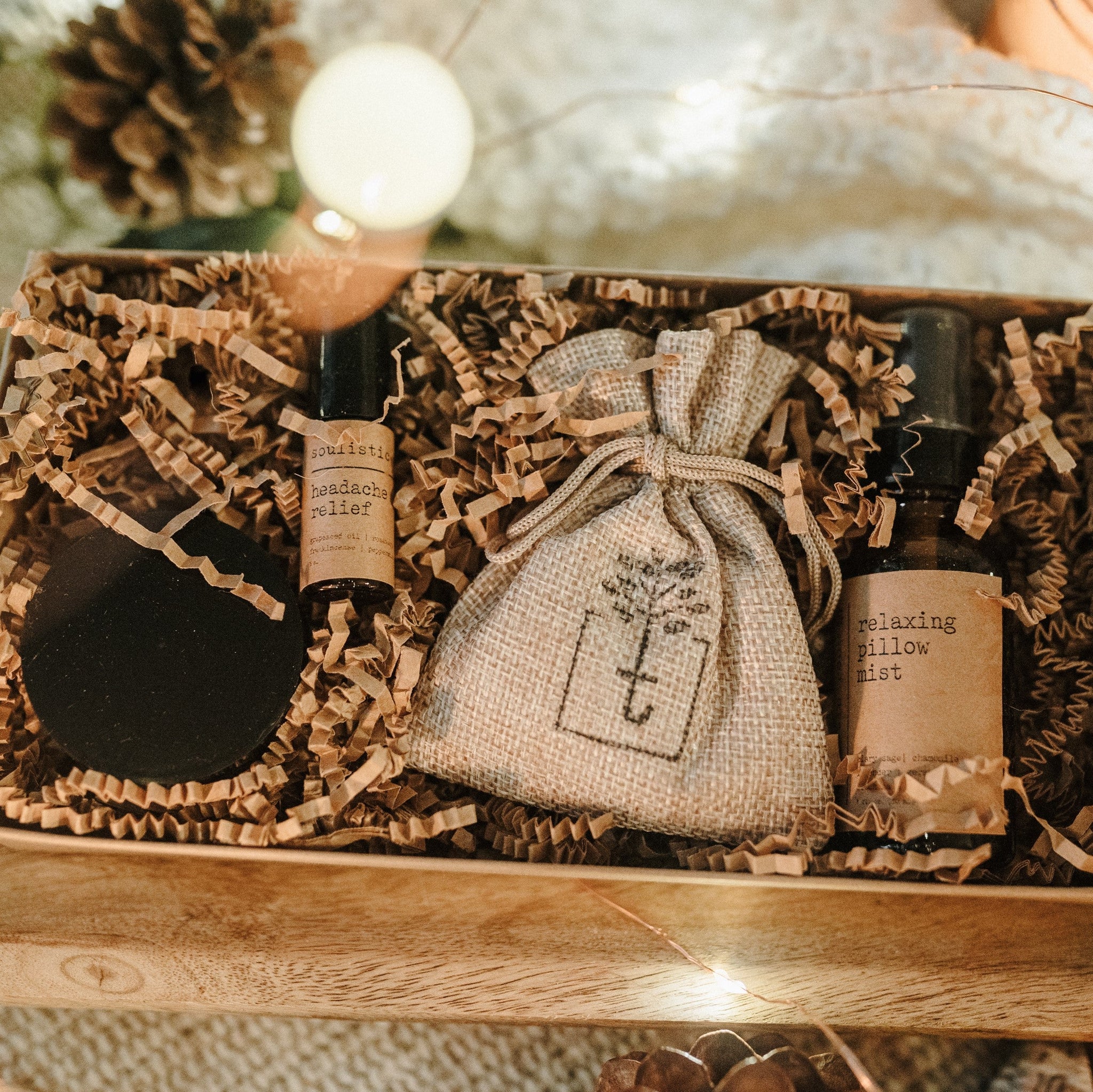 A travel set full of sample sizes already wrapped in a kraft box. The set includes a lavender & chamomile epsom salt bath, essential oil headache relief roller, organic herbal oatmeal bath soak & a relaxing pillow spray. The set is on a piece of wood and blanket with pine cones and lights in the background.